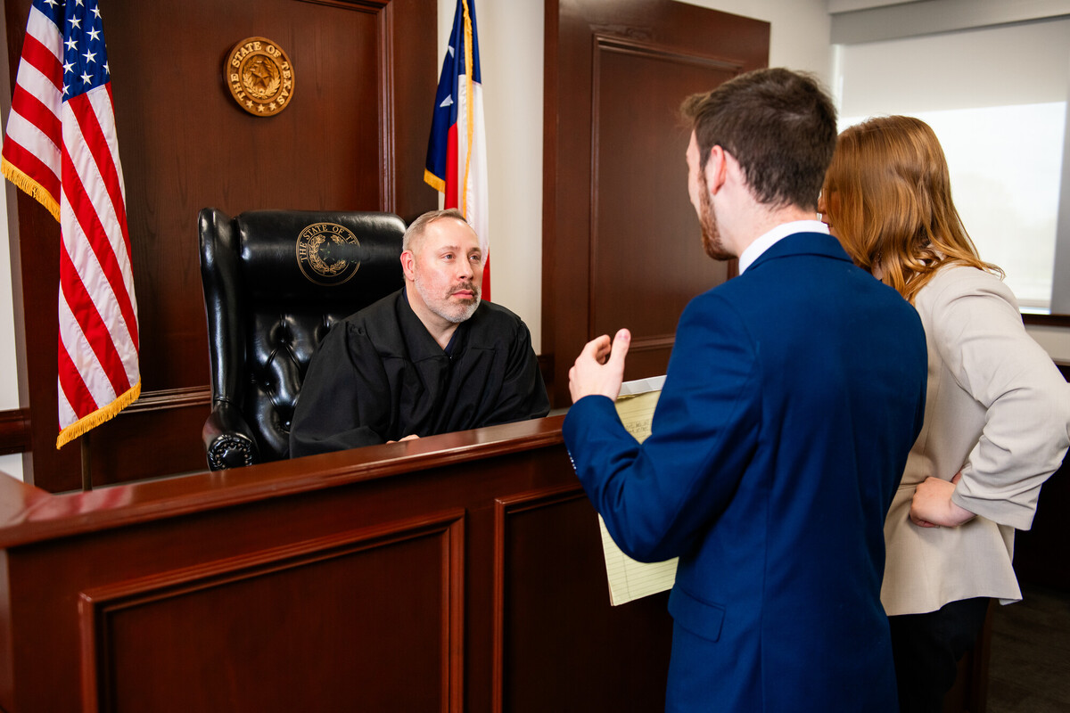 Judge at bench listens as two Collin College students present arguments during a mock trial, U.S. and Texas flags behind.