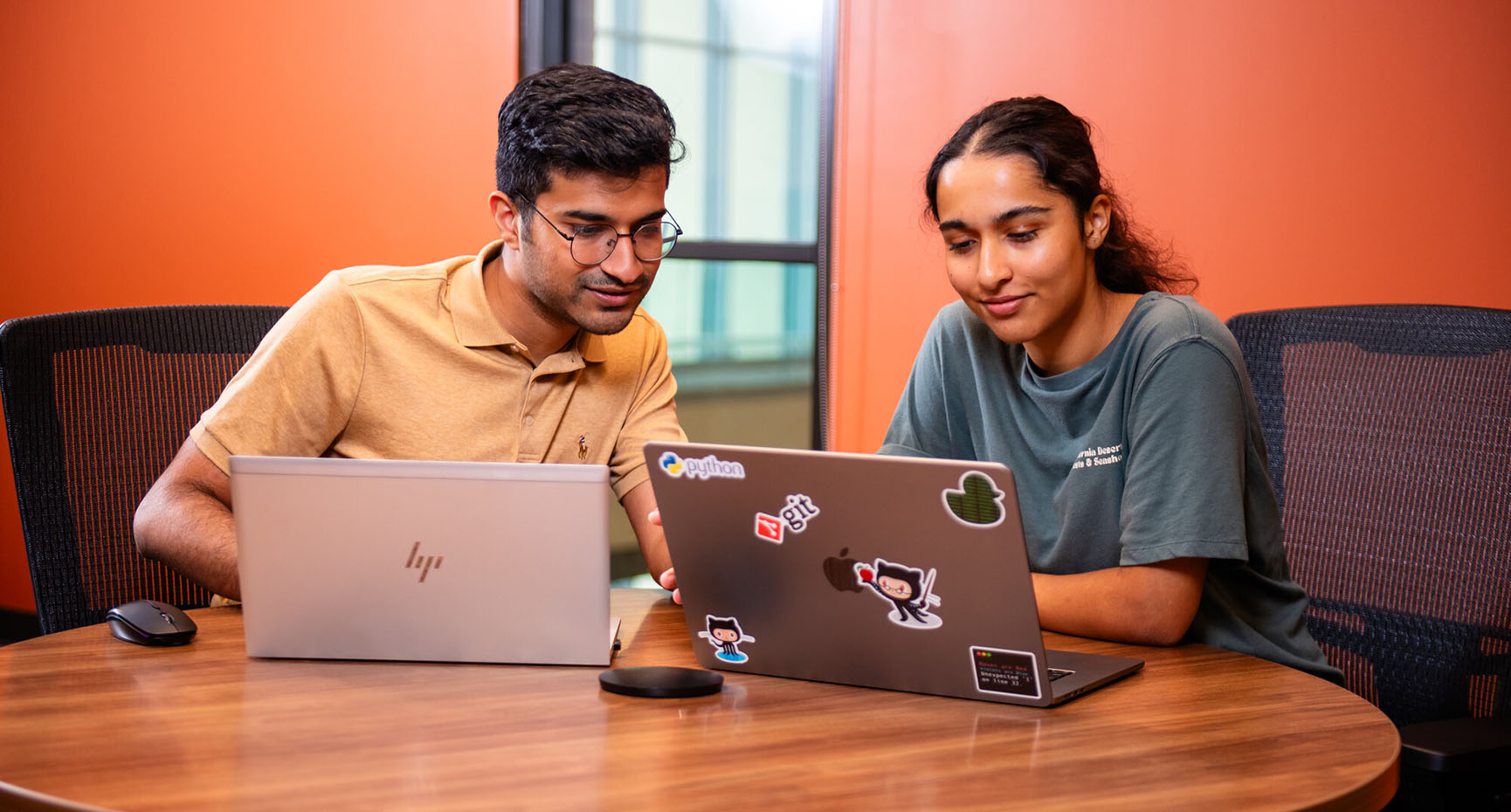Two students look at a laptop on a table. 